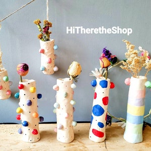 May include: Six colorful ceramic vases with various patterns and textures, including polka dots, stripes, and abstract designs. The vases are hanging or standing on a wooden surface and are filled with dried flowers.