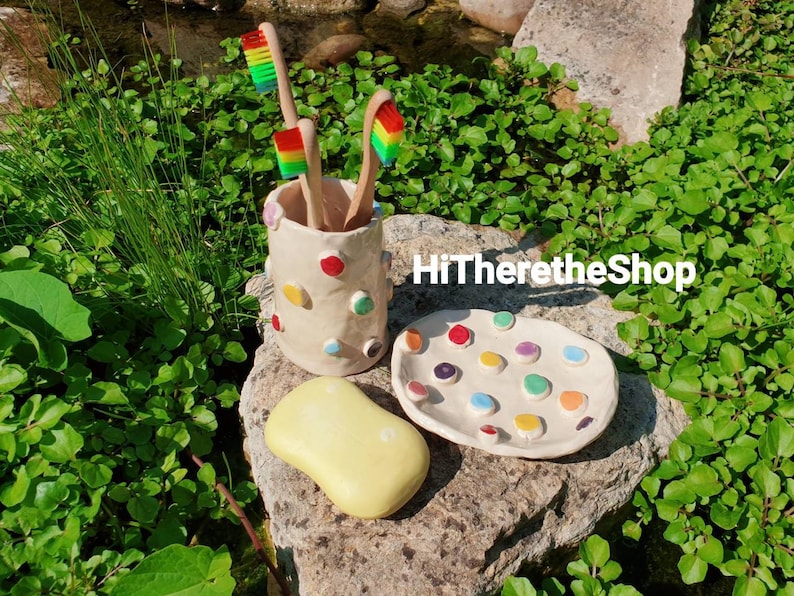 May include: A white ceramic toothbrush holder with colorful polka dots, three rainbow toothbrushes, and a white ceramic dish with colorful dots. The items are on a rock surface with green plants in the background.