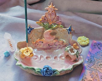 May include: A ceramic incense burner tray in a pale pink hue, featuring a sculpted central moon, a star-topped floral element, and a blue rose at the base. The tray includes a small yellow flower incense holder and a moon-shaped candle holder.