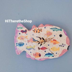 May include: A white ceramic clock shaped like a fish with a pink outline. The clock face is decorated with colourful painted fish and bubbles. The clock hands are gold.