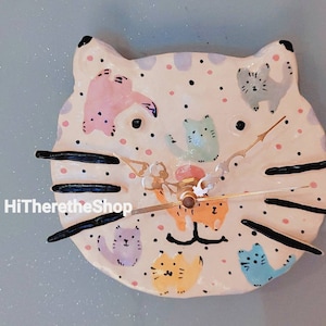 Puede incluir: Un reloj de cerámica con forma de gato con un patrón de lunares rosa, blanco y negro. La esfera del reloj presenta una variedad de gatos coloridos y una aguja de reloj dorada.