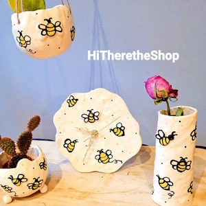 May include: A set of four white ceramic planters with a bee design. The planters are decorated with black bees and yellow accents. The planters are in various shapes and sizes, including a hanging planter, a vase, a clock, and a small pot with legs. The planters are on a wooden surface. HiThereTheShop is written on the wall behind the planters.