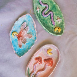Könnte beinhalten: Drei handgefertigte Keramik-Schmuckschalen. Eine ist blau mit einer rosa Blume, eine andere ist grün mit einer lila Schlange, und die letzte ist rosa mit zwei orangefarbenen Pilzen. Jede Schale hat einen gelben Akzent.