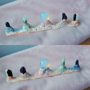 Pode incluir: Um suporte de cerâmica branca com um design ondulado, apresentando cinco pontas de cristal e duas esferas iridescentes. O suporte é decorado com pontos dourados e tem um esquema de cores azul claro, rosa e verde.