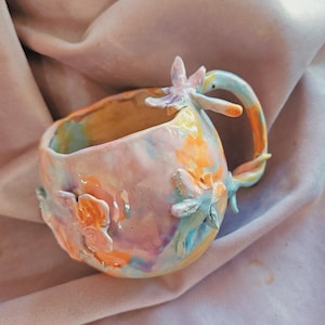 May include: A handmade ceramic mug with a rounded, irregular shape, featuring a pastel swirl of pink, orange, yellow, and blue glaze. The mug is decorated with three-dimensional sculpted flower appliqués and has a curved handle.