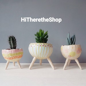 May include: Three small ceramic planters with three-legged bases. Each planter is painted with a different pattern in pink, yellow, and green. The planters are filled with small plants.