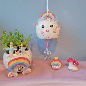 Op de afbeelding: Een set van vier keramische potten met een regenboogthema. De potten zijn versierd met grillige figuren en kleurrijke regenbogen. Eén pot is een hangende pot in de vorm van een wolk met een glimlachend gezicht. Een andere pot is een grotere pot met een regenboog en twee vogels. De overige twee potten zijn kleiner en in de vorm van paddenstoelen.