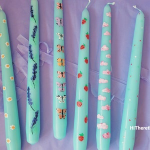 May include: Six teal taper candles with different painted designs. The designs include daisies, lavender, butterflies, strawberries, clouds, and polka dots.