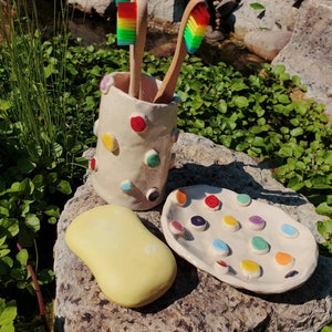 May include: A white ceramic toothbrush holder with colorful polka dots, a yellow soap dish, and a white ceramic dish with colorful dots. The holder has two rainbow toothbrushes inside.