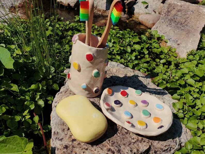May include: A white ceramic toothbrush holder with colorful polka dots, a yellow soap dish, and a white ceramic dish with colorful dots. The holder has two rainbow toothbrushes inside.
