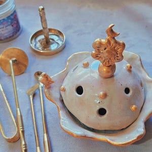Peut inclure: Un brûleur d'encens en céramique avec un couvercle en dôme crème orné d'accents dorés, d'un fleuron soleil et lune, et de petites sphères dorées. L'ensemble comprend des outils en métal doré, un petit support circulaire et un pot d'encens.