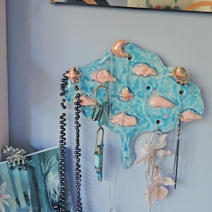 May include: A whimsical, cloud-shaped ceramic jewelry holder in shades of blue and pink, adorned with a crescent moon and various hanging necklaces and earrings. The holder is mounted on a light blue wall, creating a decorative and functional display.