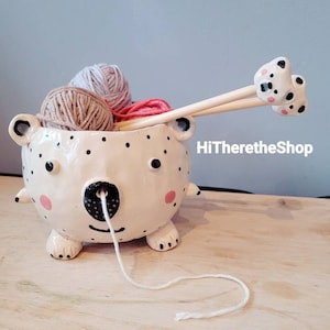 May include: White ceramic yarn bowl shaped like a bear with black polka dots, pink cheeks, and a black nose. The bowl has a hole in the nose for the yarn to feed through. Two wooden knitting needles with bear head handles are in the bowl. The bowl is filled with three balls of yarn: beige, light pink, and light gray.