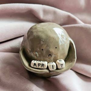 Puede incluir: Una figurita de cerámica gris con una cara triste, sosteniendo tres pequeños bloques blancos con las palabras "MAR", "APR" y "01" escritas en ellos.