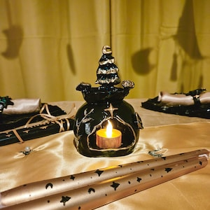 Könnte beinhalten: Ein schwarzer Kerzenhalter aus Keramik mit einem goldenen Baumdesign und einer kleinen Kerze, die darin brennt. Der Halter steht auf einer goldenen Stoffoberfläche mit zwei goldenen Kerzenständern mit schwarzen Sternen- und Halbmondmustern.