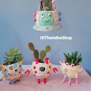 Peut inclure: Trois pots en céramique en forme de personnages de dessins animés avec des plantes à l'intérieur. Les pots sont peints dans des couleurs vives avec des détails comme des yeux, des bouches et des points. Les pots sont posés sur une surface rose pâle. HiTheretheShop est écrit en noir sur fond blanc.
