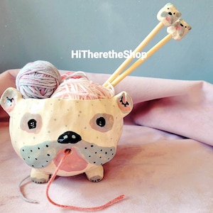 May include: A whimsical, hand-painted ceramic yarn bowl shaped like a bulldog. The bowl is cream-colored with black and gray accents, holding pink and gray yarn balls. Two wooden knitting needles with bear-shaped toppers rest inside. The text "HiTheretheShop" is visible.