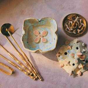 Peut inclure: Un ensemble d'outils en métal doré comprenant des pinces, une brosse et des ustensiles à long manche, accompagnés d'un petit bol en céramique à motif floral, d'un porte-encens circulaire en métal et d'un couvercle en céramique texturé.
