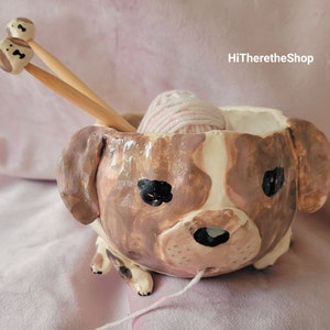 Könnte beinhalten: Keramische Schale in Form eines Hundes mit einer rosa Wollknäuel im Inneren und zwei Holzstricknadeln mit braun-weiß bemalten Spitzen, die aus der Schale herausragen. Die Schale ist braun-weiß bemalt, um einem Hundegesicht zu ähneln. HiThereTheShop ist im Hintergrund geschrieben.
