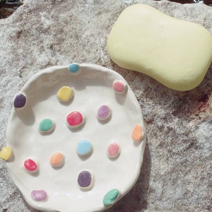 May include: A white ceramic dish with a scalloped edge and colorful dots. The dish is on a light gray stone surface. A yellow bar of soap is next to the dish.