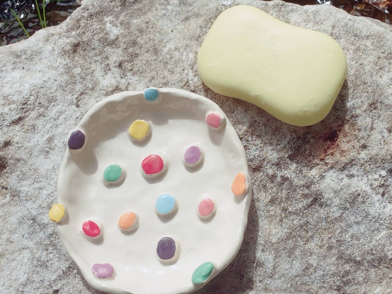 May include: A white ceramic dish with a scalloped edge and colorful dots. The dish is on a light gray stone surface. A yellow bar of soap is next to the dish.