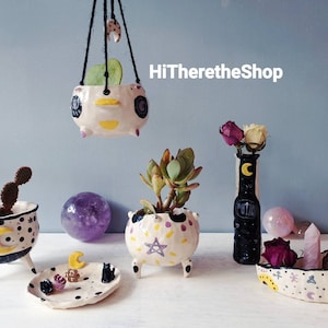 Könnte beinhalten: Eine Sammlung handgefertigter Keramik-Blumentöpfe und Vasen mit skurrilen Designs mit Mond, Sternen und anderen himmlischen Motiven. Die Töpfe sind mit schwarzen, gelben und violetten Akzenten verziert. HiThereTheShop ist im Bild geschrieben.