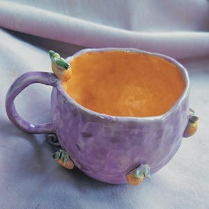 May include: A handmade ceramic mug with a lavender exterior and an orange interior. The mug features a handle and is adorned with small bird-like embellishments in green and orange. The mug is a unique piece of art.