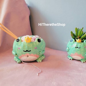 May include: Two green frog-shaped ceramic planters with gold crowns. One planter has a pink interior and is filled with yarn and a wooden knitting needle. The other planter has a succulent plant growing inside. The text "HiThereTheShop" is visible in the background.