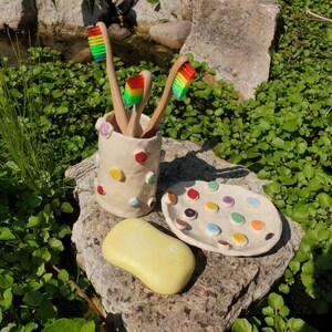 May include: A white ceramic toothbrush holder with colorful polka dots, holding four toothbrushes. The holder is sitting on a rock with a white ceramic dish with colorful dots. A yellow bar of soap is also on the rock.