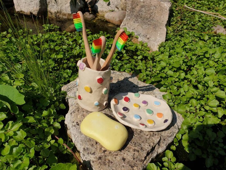 May include: A white ceramic toothbrush holder with colorful polka dots, holding four toothbrushes. The holder is sitting on a rock with a white ceramic dish with colorful dots. A yellow bar of soap is also on the rock.