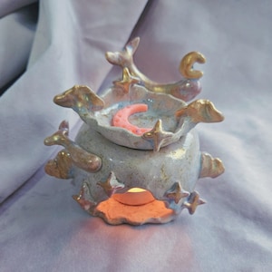 Puede incluir: Un quemador de aceite de cerámica blanca con un diseño de luna y estrellas. El quemador tiene un pequeño plato en la parte superior para contener aceites esenciales o ceras derretidas. El quemador está encendido y brillando.