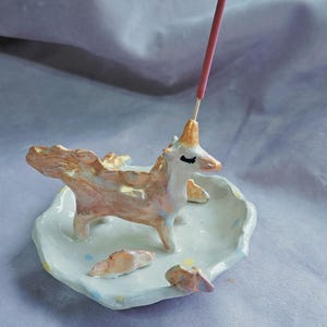 May include: A ceramic unicorn incense holder with a pink incense stick. The unicorn is white with gold accents and stands on a light blue dish. The dish has small, decorative, tan-coloured rocks.