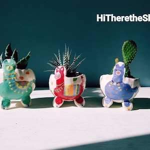 May include: Three ceramic planters shaped like llamas, each with a different colour and design. The planters are filled with succulents and are sitting on a white surface. HiThereTheShop is written in white text on a blue background.