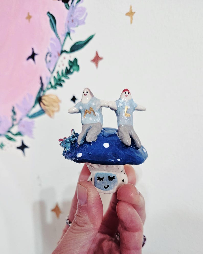 K&ouml;nnte beinhalten: Zwei kleine Keramikfiguren mit blauer und wei&szlig;er Kleidung stehen auf einem blauen und wei&szlig;en Pilz mit einem l&auml;chelnden Gesicht. Die Figuren halten sich an den H&auml;nden und haben die Buchstaben "M" und "L" auf ihren Hemden.