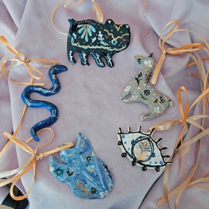 May include: Five ceramic ornaments with gold and blue paint details. The ornaments are shaped like a snake, a hippopotamus, a deer, an eye, and a hand. Each ornament has a gold ribbon attached.