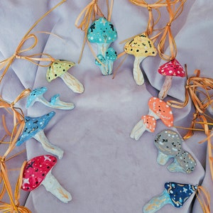 Puede incluir: Una colección de 12 adornos de cerámica con forma de hongo pintados a mano con varios colores y patrones. Cada adorno tiene una cuerda dorada para colgar. Los hongos se muestran sobre un fondo de tela de color púrpura claro.