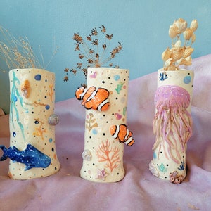 Può includere: Tre vasi in ceramica con disegni a tema oceanico. Il vaso a sinistra ha uno squalo blu, il vaso al centro ha pesci pagliaccio arancioni e bianchi, e il vaso a destra ha una medusa rosa.