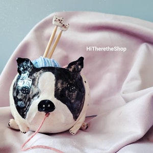 May include: A ceramic yarn bowl shaped like a black and white Boston Terrier dog with a blue and white ball of yarn inside and two wooden knitting needles sticking out. The dog is sitting on a pink fabric surface with a pink yarn tail trailing behind it.