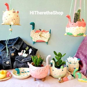 Pode incluir: Uma coleção de vasos e decorações de dinossauros de cerâmica feitos à mão. Os vasos são pintados em uma variedade de cores, incluindo rosa, verde e amarelo. As decorações incluem um sino de vento de dinossauro pendurado, um relógio de dinossauro e um pequeno prato de dinossauro. O texto "HiThereTheShop" é visível na imagem.