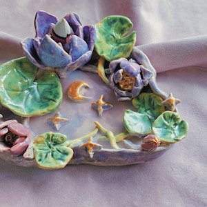 May include: A ceramic incense burner shaped like a pond with water lilies, a crescent moon, and stars. The lilies are painted in shades of purple, green, and gold.