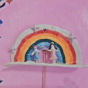 May include: A semi-circular ceramic wall hanging featuring a hand-painted rainbow with two small figures standing on a ledge. The rainbow is colored in red, orange, yellow, green, and blue, accented with three-dimensional star shapes and a gold chain.
