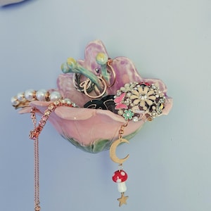 以下が含まれることがあります： ピンクと紫のセラミック製の花型のジュエリーホルダー。リング、ブレスレット、イヤリングなど、様々なジュエリーが収められています。ホルダーは水色の壁に取り付けられています。キノコと月のチャームがぶら下がっています。寸法はセンチメートルです。