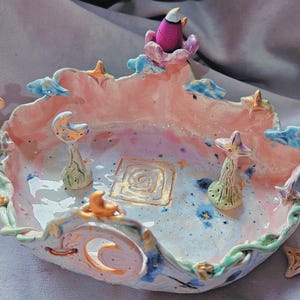 May include: A ceramic bowl with a pastel pink and blue glaze, featuring a central gold square spiral, two small mushroom-like figures, and a crescent moon cutout. The rim is decorated with small stars and clouds, accompanied by a separate star-shaped stand.