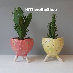 Peut inclure: Deux pots de fleurs en céramique avec trois pieds chacun. Le pot de gauche est rose avec des pois blancs et une plante de cactus. Le pot de droite est jaune avec des pois blancs et orange et une plante de cactus verte. HiThereTheShop est écrit en blanc sur un fond gris.