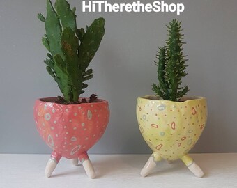 May include: Two ceramic plant pots with three legs each. The pot on the left is pink with white polka dots and a cactus plant. The pot on the right is yellow with white and orange polka dots and a green cactus plant. HiThereTheShop is written in white text on a gray background.