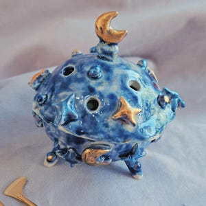 Puede incluir: Un quemador de incienso de cerámica azul de forma redonda con esmalte moteado, decorado con estrellas y lunas crecientes doradas en relieve. La tapa tiene orificios de ventilación y un asa en forma de luna creciente.