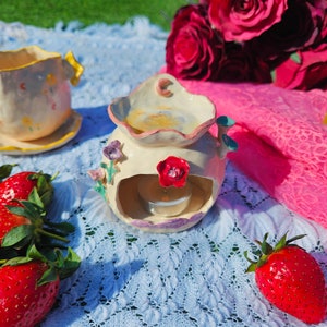 Könnte beinhalten: Eine weiße Teetasse und Untertasse aus Keramik mit Blumenmuster, ein weißer Keramik-Tee-Wärmer mit Blumenmuster und einer rosa Rose sowie ein Bund roter Erdbeeren auf einer weißen Häkeltischdecke.