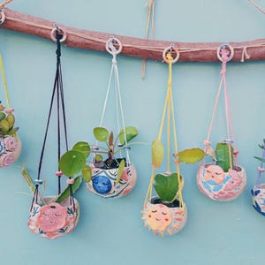 May include: Six colorful ceramic hanging planters with various plants, each planter features a different hand-painted face with a whimsical expression. The planters are suspended from a wooden branch with macrame string.