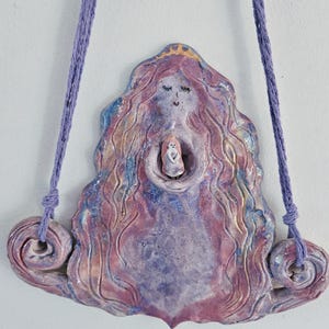May include: A ceramic wall hanging depicting a woman with long, flowing hair. The woman is holding a smaller figure in her arms. The wall hanging is painted in shades of purple, pink, and blue.