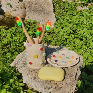 May include: A white ceramic toothbrush holder with colorful polka dots, holding four rainbow-colored toothbrushes. A matching white ceramic dish with colorful polka dots is next to the holder, and a yellow bar of soap is in front of the dish.